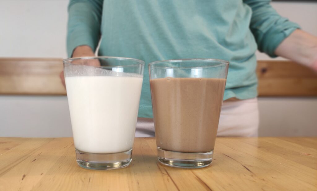 un vaso con leche de amaranto blanca y otro vaso con leche de amaranto y cacao de color chocolate reposan sobre una mesa de madera