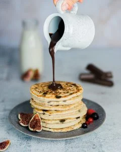 torre de pancakes sobre un plato y sobre ella, una jarra que vierte chocolate por encima de las tortitas
