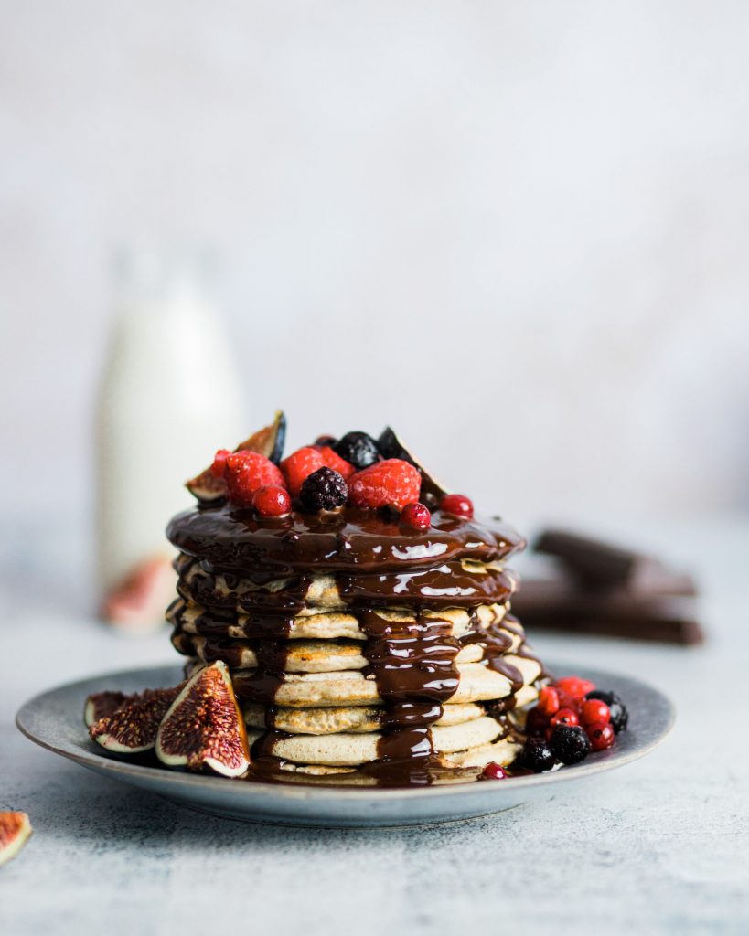 torre hecha con 8 pancakes sobre un plato. Llevan un baño de chocolate y frutas del bosque como topping