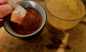 chocolate a la taza junto a un vaso con grano de avena a remojo y una mano untando bizcocho en el chocolate