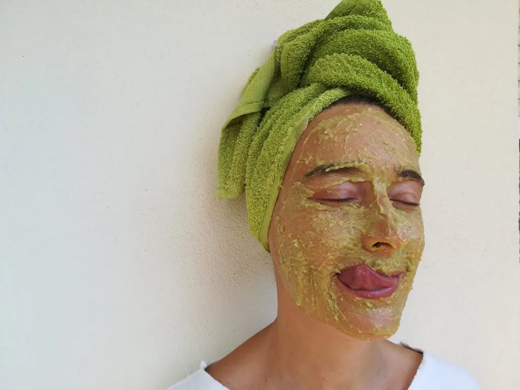 cara de persona con una mascarilla color aguacate y toalla verde en la cabeza sobre fondo blanco. La persona saca la lengua chupando la mascarilla facial